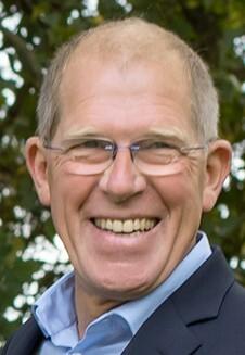 Näher der lächelnde Mann mit Brille, mittellangen grauen Haaren und blazer, vor einem unscharfen grünen Hintergrund.