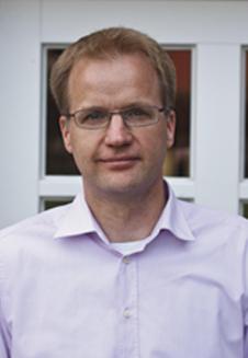 Porträt eines Mannes mit Brille und blauer Hemd vor einem Fenster mit weißen Rahmen.
