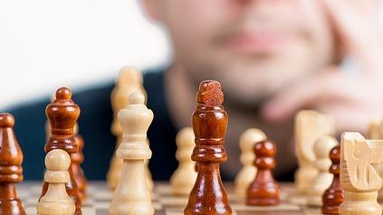 Nahaufnahme eines Schachbretts mit Holzfiguren, im Hintergrund ein nachdenklicher Mann mit Brille.