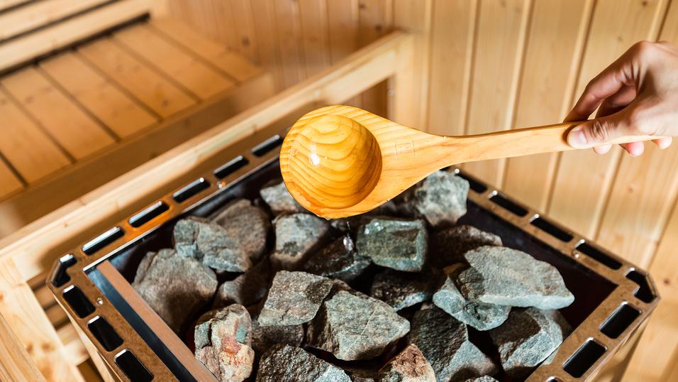 Holzlöffel mit Wasser über einem heißen Steinkessel in einer Sauna. Sauna-Holzwände im Hintergrund sichtbar.