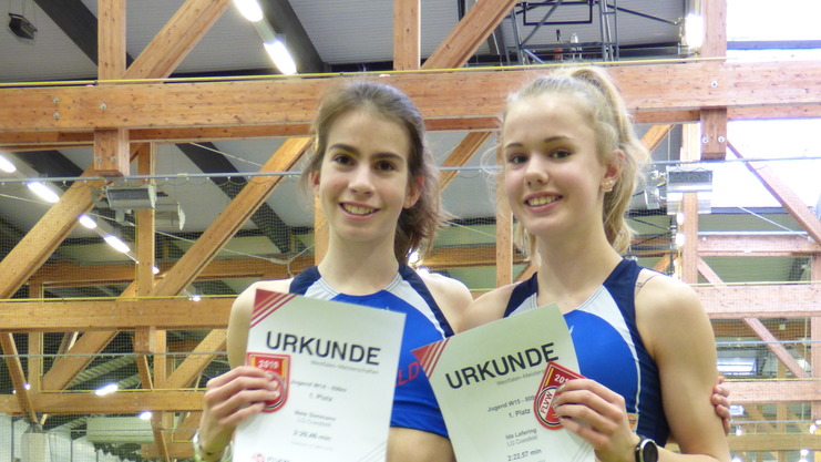 Zwei junge Athletinnen in Sportbekleidung halten Urkunden für ihre Platzierungen bei einem Leichtathletikwettbewerb.