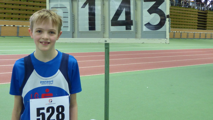 Junger Sportler in blauer Kleidung steht neben einer Wettkampfanzeige mit den Nummern 528 und 143 in einer Sporthalle.