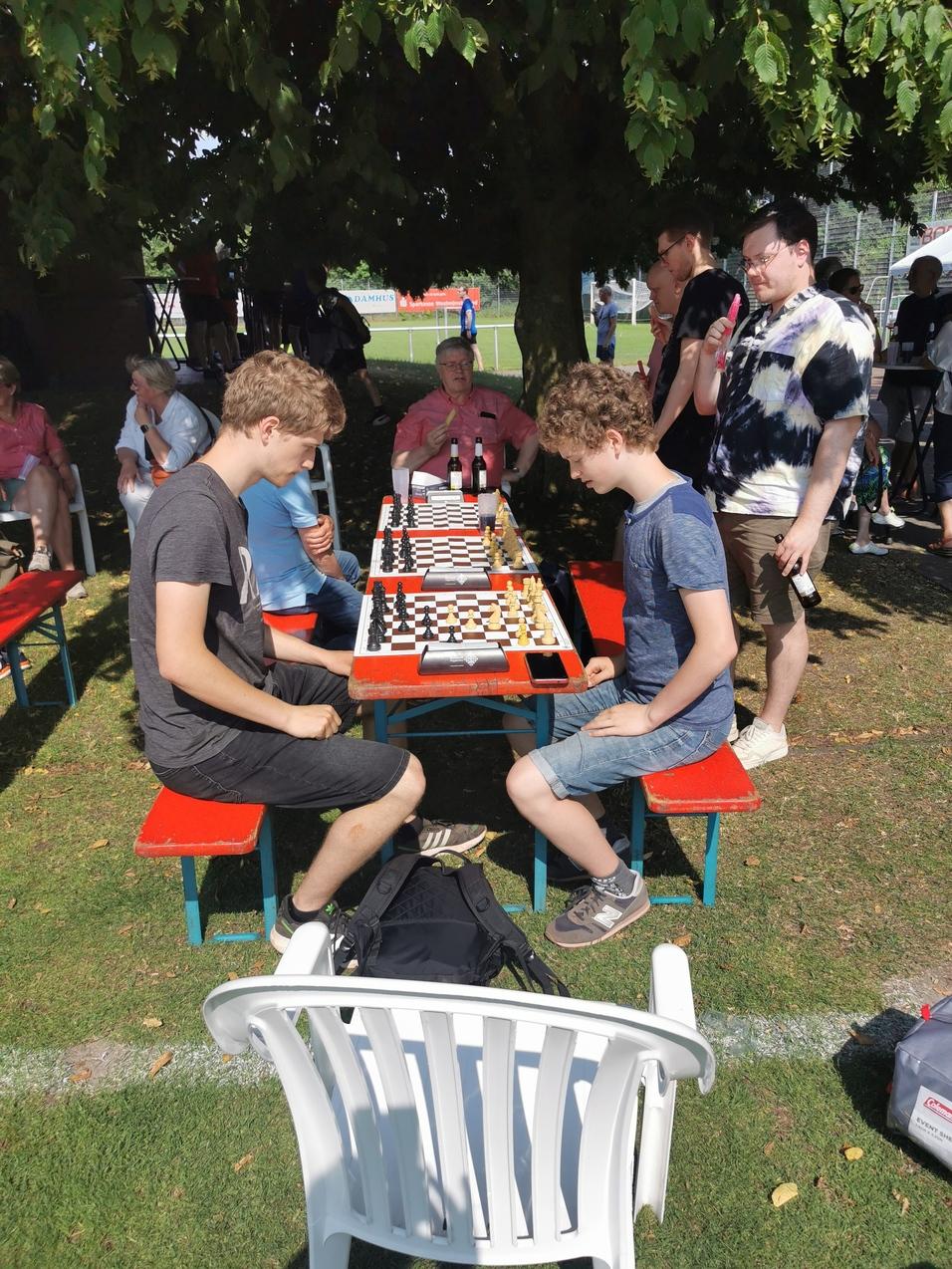 Zwei Jugendliche spielen Schach an einem Tisch im Freien, umgeben von Zuschauern und Bänken unter einem Baum.