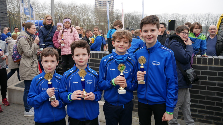 Vier Jungen in blauen Sportjacken halten Pokale vor einer Menschenmenge in einem städtischen Umfeld.