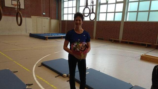 Frau steht in einer Turnhalle mit einem Blumenstrauß in der Hand, Matten und Turngeräte im Hintergrund.
