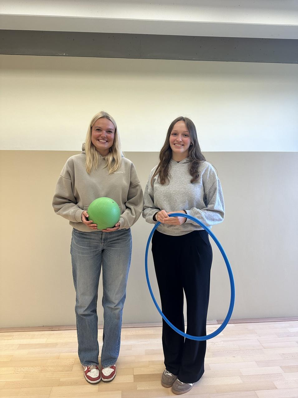 Zwei Frauen stehen in einem Raum, eine hält einen grünen Ball, die andere einen blauen Hula-Hoop-Reifen.