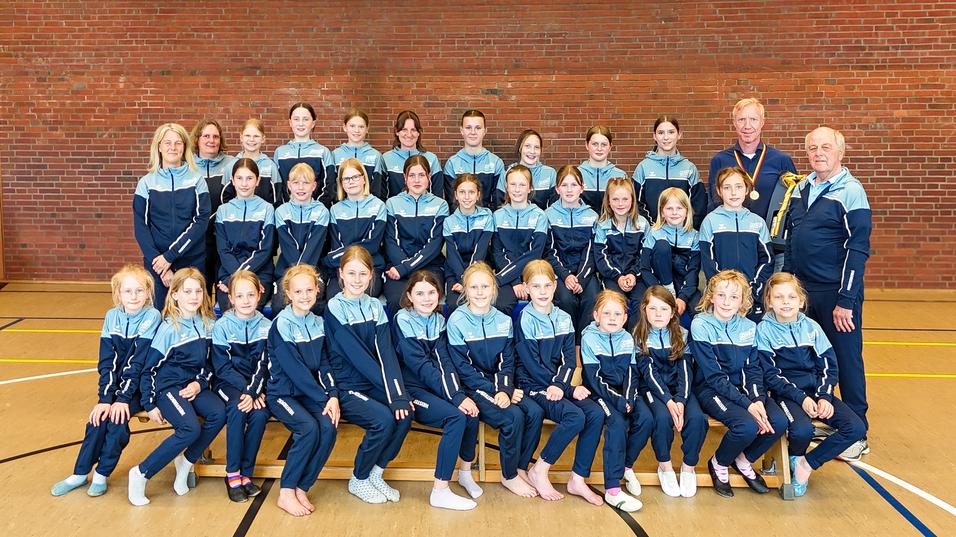 Gruppenfoto von 31 Mädchen und Betreuern in Sportanzügen, aufgestellt vor einer Ziegelsteinwand in einer Sporthalle.
