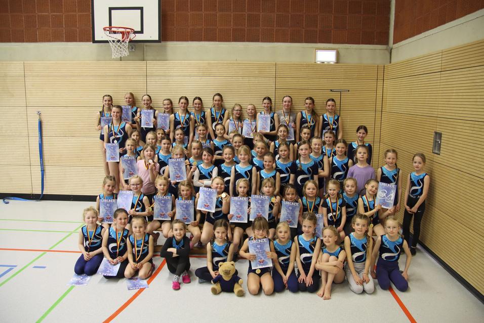 Gruppenfoto von 60 Gymnastinnen in blauen Trikots mit Medaillen und Urkunden, auf einem Sportplatz in der Halle.