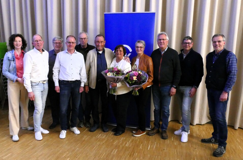 Gruppe von neun Personen steht lächelnd vor einem Banner, einige halten Blumensträuße in den Händen.