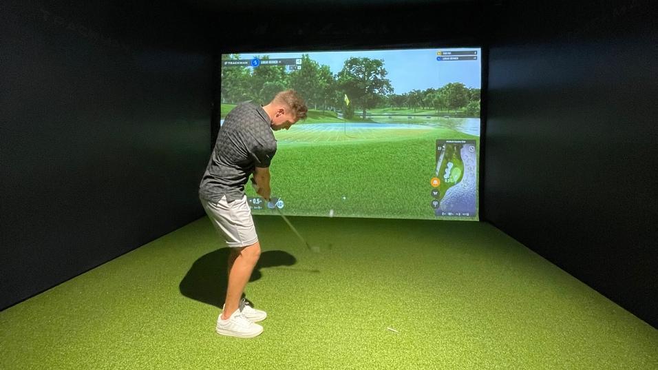 Mann schl&auml;gt mit einem Golfschl&auml;ger in einem Simulatorraum mit gr&uuml;nem Kunstrasen und Bildschirmhintergrund.