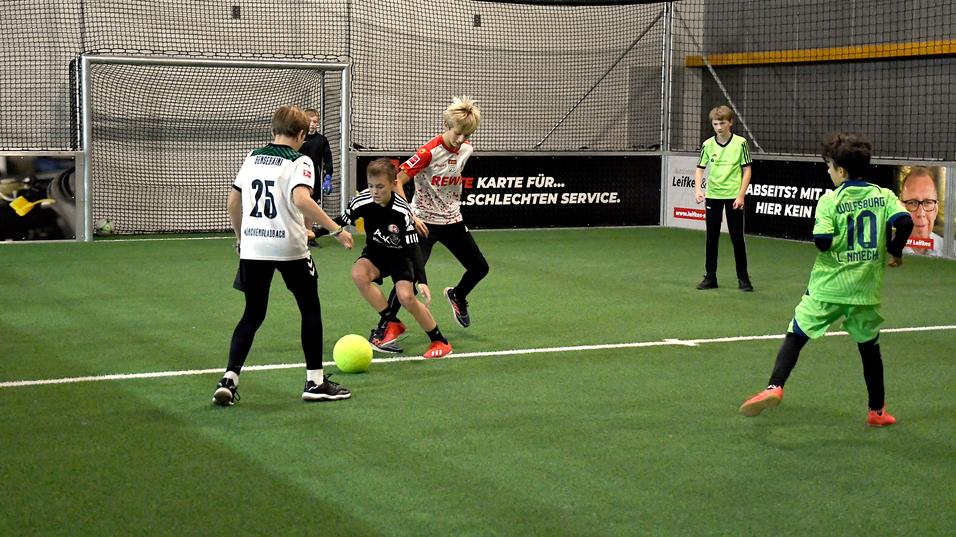 Vier Kinder spielen Fußball in einem Innenraum mit grünem Kunstrasen und einem Tor im Hintergrund.