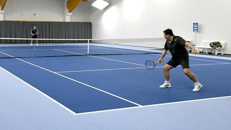 Zwei Tennisspieler in einer Halle, einer schlägt den Ball mit einem Schläger, blauer Boden und Netz im Hintergrund sichtbar.