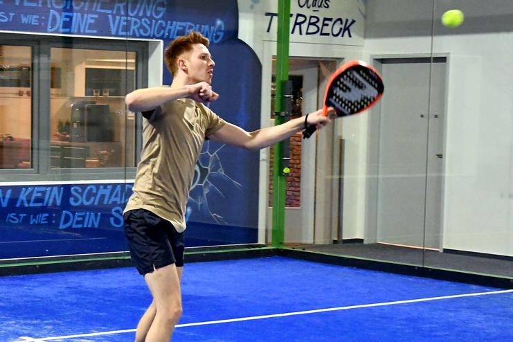 Junger Spieler schlägt mit einem Paddel einen Ball auf einem blauen Padelplatz mit Wänden im Hintergrund.