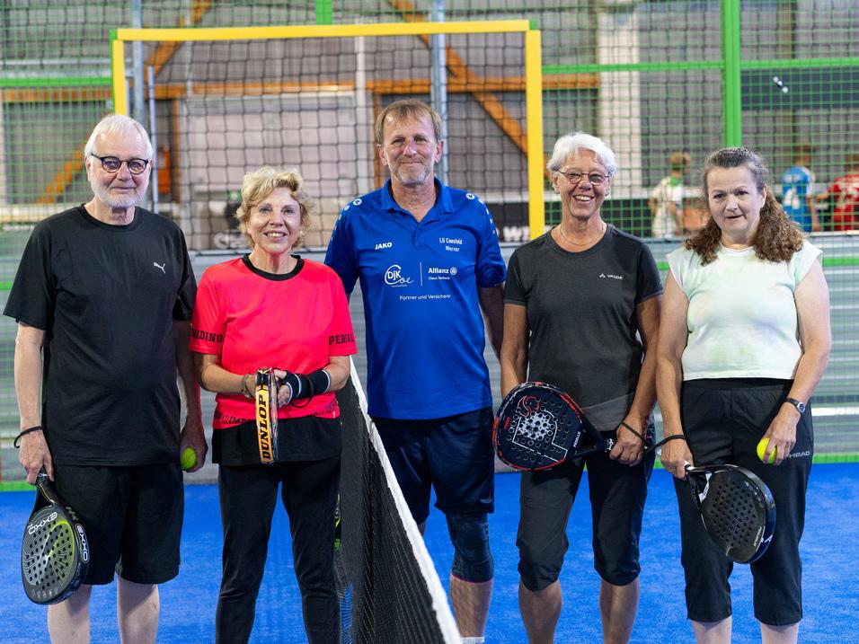 Gruppe von fünf Padelspielern posiert auf einem Padelplatz, jeder hält Schläger und einen Ball in der Hand.