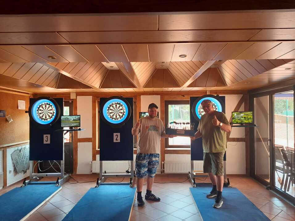 Zwei Männer stehen in einem Dartsalon vor Dartscheiben und präsentieren einen Pokal zwischen ihnen.