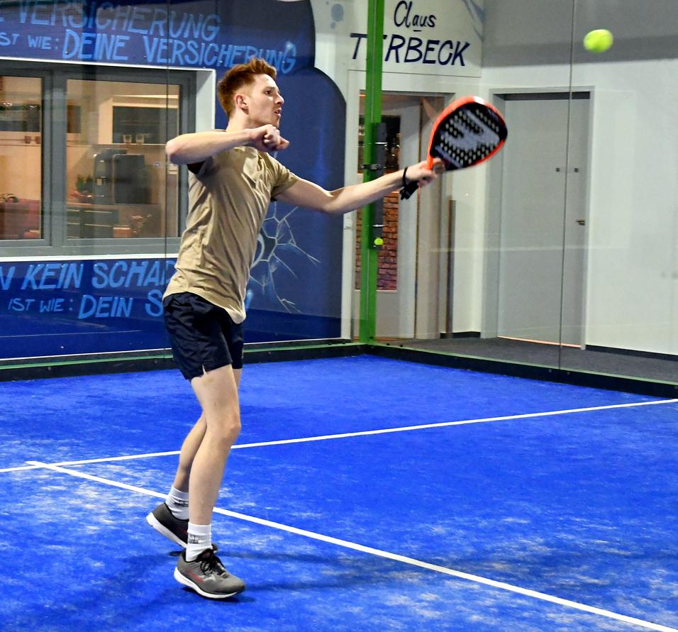 Junger Spieler schlägt mit einem Paddel einen Ball auf einem blauen Padelplatz mit Wänden im Hintergrund.