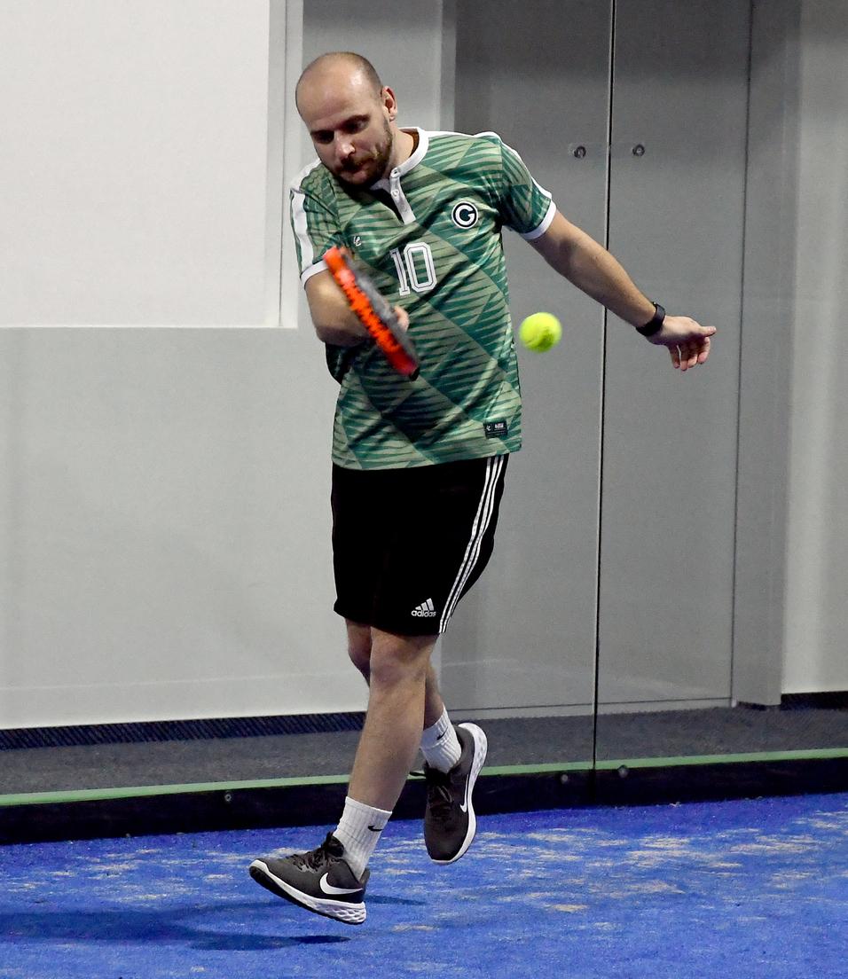 Mann in sportlicher Kleidung schlägt einen Tennisball mit einem Schläger in einer Halle mit blauen Bodenbelag.