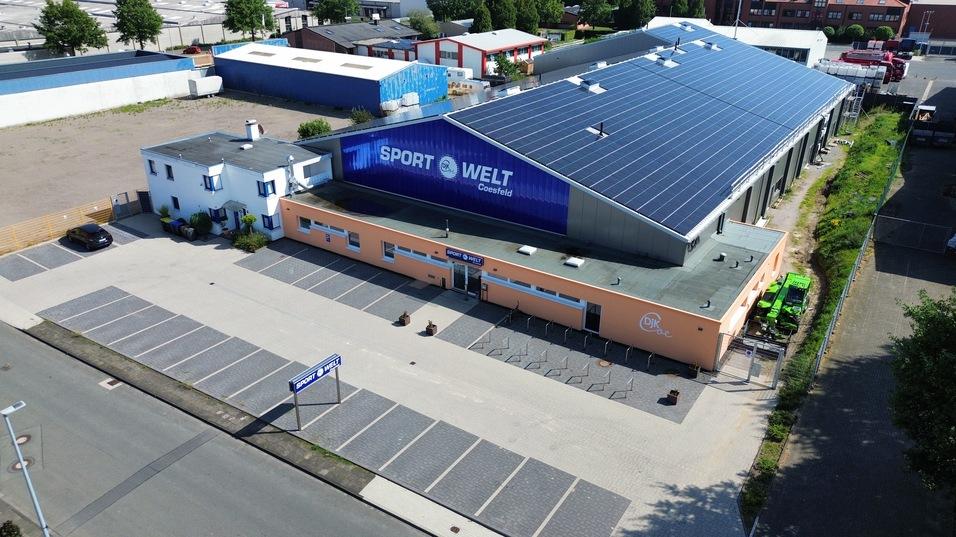 Luftaufnahme eines Sportzentrums mit Parkplatz, blauer Fassade und Solaranlagen auf dem Dach.