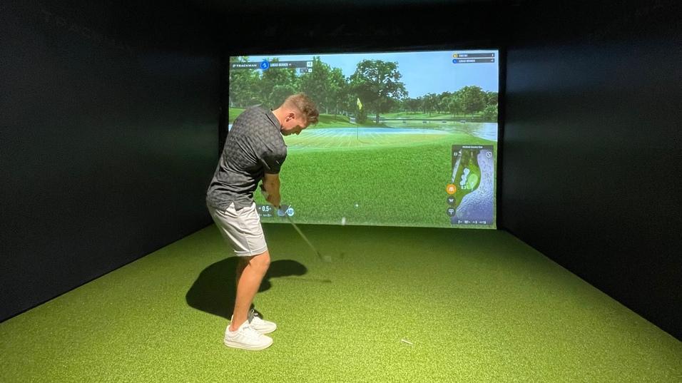 Mann schlägt mit einem Golfschläger in einem Simulatorraum mit grünem Kunstrasen und Bildschirmhintergrund.