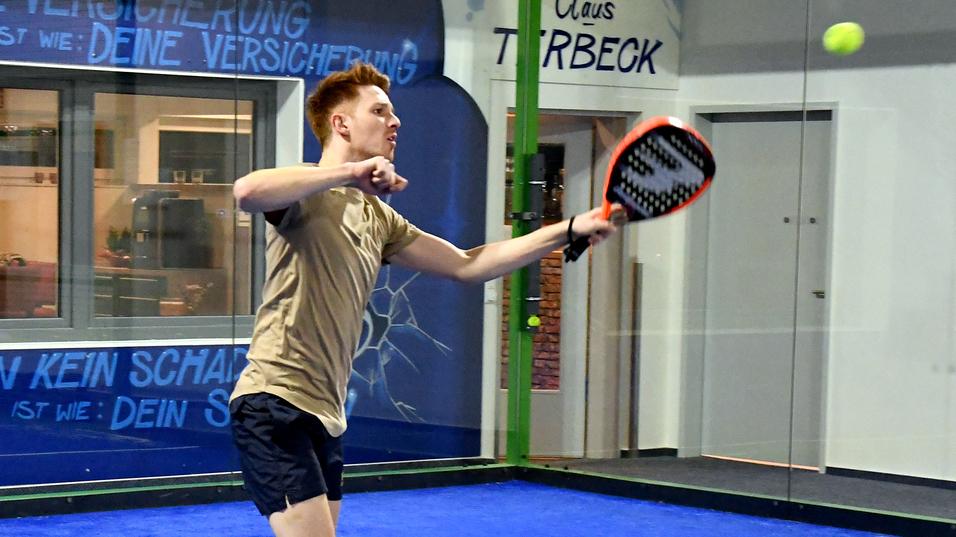 Junger Spieler schl&auml;gt mit einem Paddel einen Ball auf einem blauen Padelplatz mit W&auml;nden im Hintergrund.