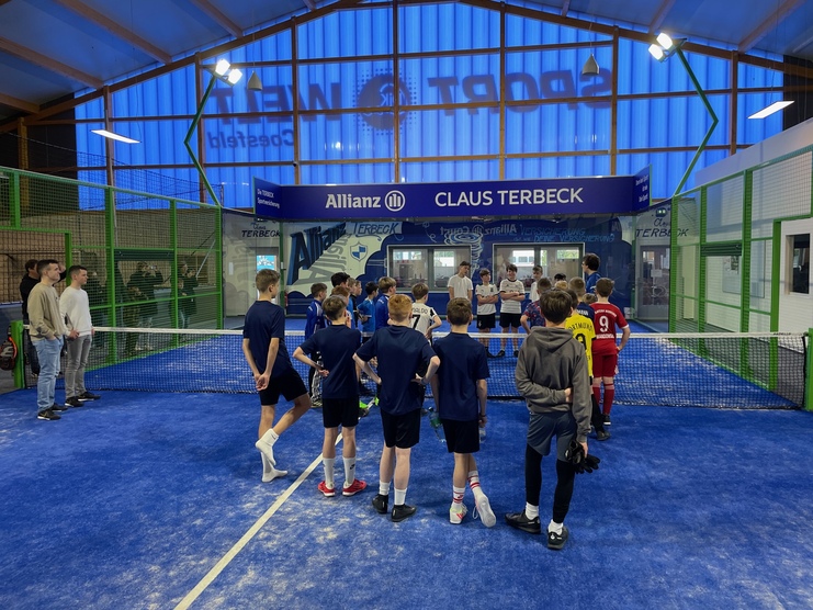 Gruppe von Jungen hört einem Trainer in einer Sporthalle mit blauem Boden und Netz zu.