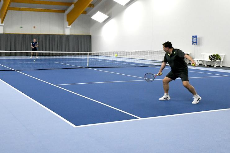 Zwei Tennisspieler in einer Halle, einer schlägt den Ball mit einem Schläger, blauer Boden und Netz im Hintergrund sichtbar.