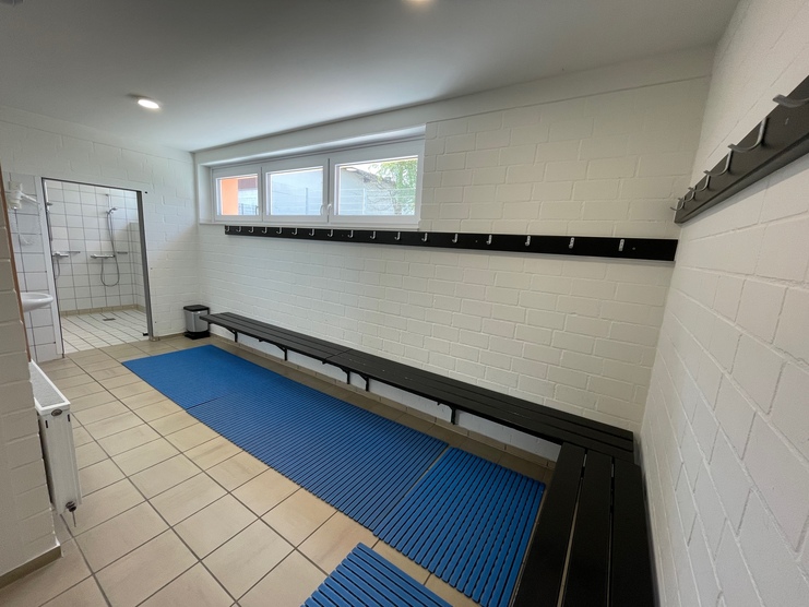 Umkleideraum mit Bänken, blauen Matten, Kleiderhaken und Dusche in hellem, modernen Ambiente.
