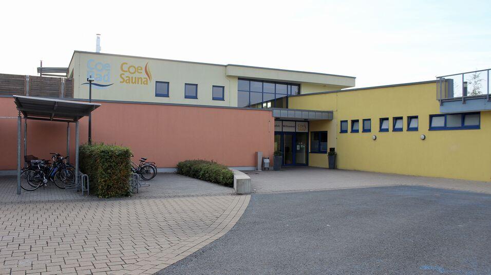 Eingang zur Coe Bad und Coe Sauna, umgeben von Fahrrädern und bepflanzten Bereichen auf einem gepflasterten Platz.