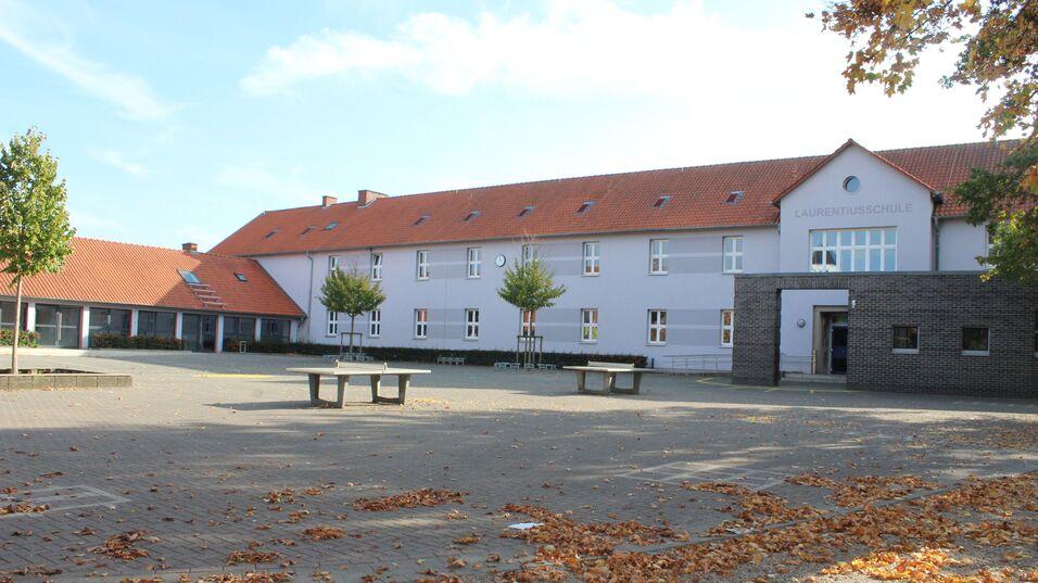 Schulgebäude mit roten Dachziegeln, umgeben von einem Pausenhof mit Laub und Bäumen im Sonnenlicht.