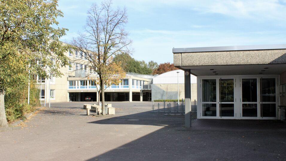 Schulgelände mit modernen Gebäuden und Bäumen, im Vordergrund der Eingang zu einem Gebäude und ein Parkplatz.