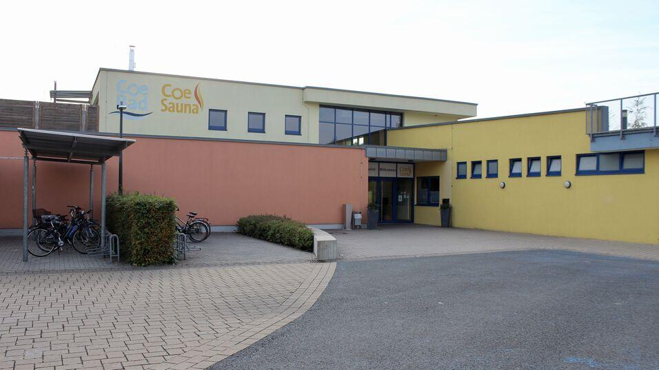 Eingang zur Coe Bad und Coe Sauna, umgeben von Fahrr&auml;dern und bepflanzten Bereichen auf einem gepflasterten Platz.