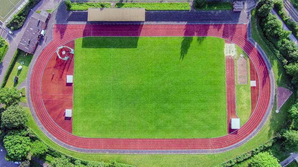Luftaufnahme eines Sportplatzes mit Laufbahn, Rasenfläche und umgeben von Bäumen und Tribünen.