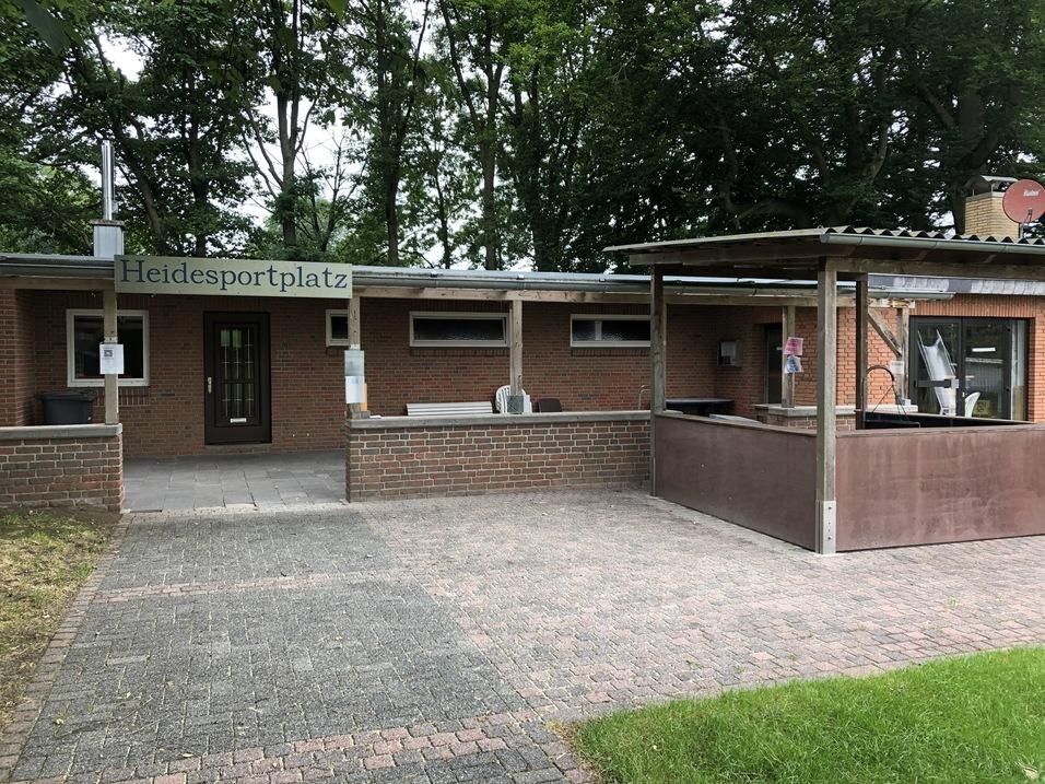 Eingang zum Heidesportplatz mit Ziegelwand, Sitzbereich und überdachter Terrasse, umgeben von Bäumen.