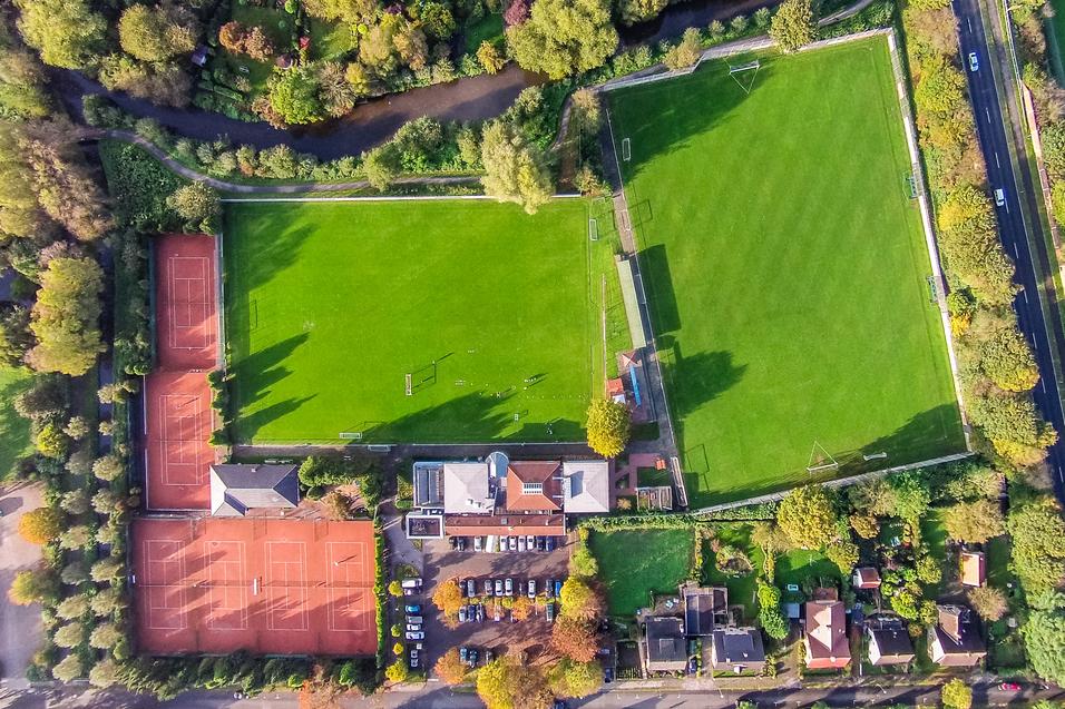 Luftaufnahme eines Sportgeländes mit zwei Tennisplätzen links und einem Fußballfeld rechts, umgeben von Bäumen.