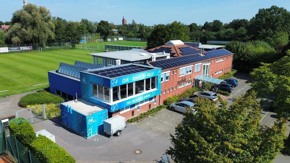 Draufsicht auf ein Sportgebäude mit Solaranlagen, umgeben von Parkplätzen, Grünflächen und einem Fußballfeld im Hintergrund.