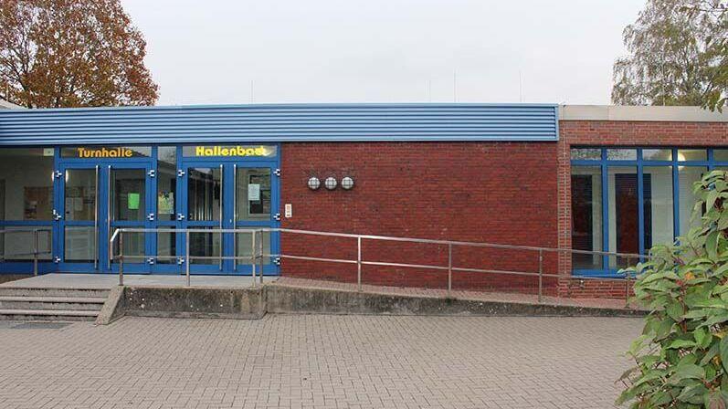Eingang zur Turnhalle mit blauen Türen und Glasfenstern, umgeben von einem gepflasterten Vorplatz und Bäumen.