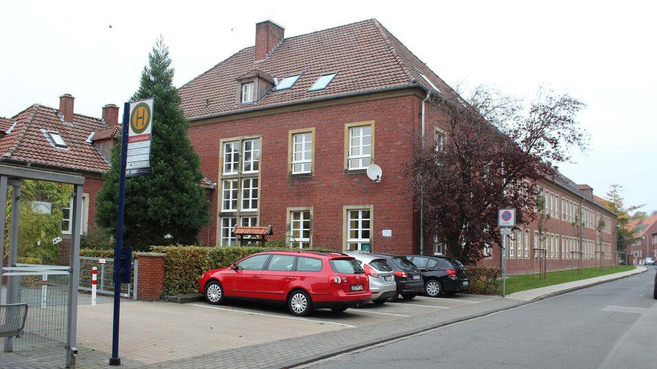 Rote und graue Autos parken vor einem zweigeschossigen Backsteingebäude mit großen Fenstern und einem Buswartehäuschen.