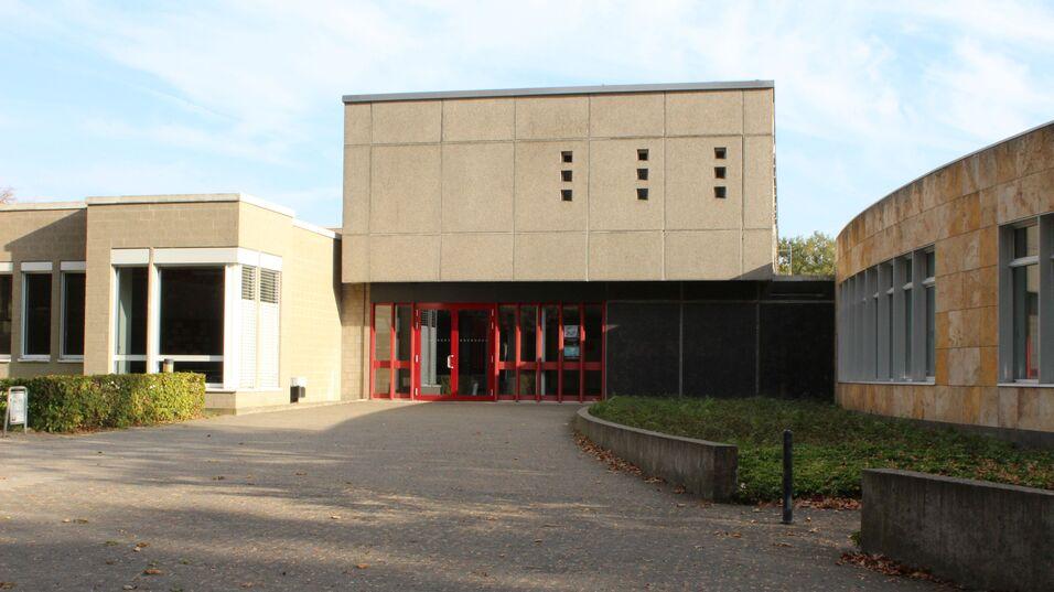 Eingang eines modernen Geb&auml;udes mit roter T&uuml;r und umliegendem Weg, flankiert von gr&uuml;nen B&uuml;schen und Steinstrukturen.