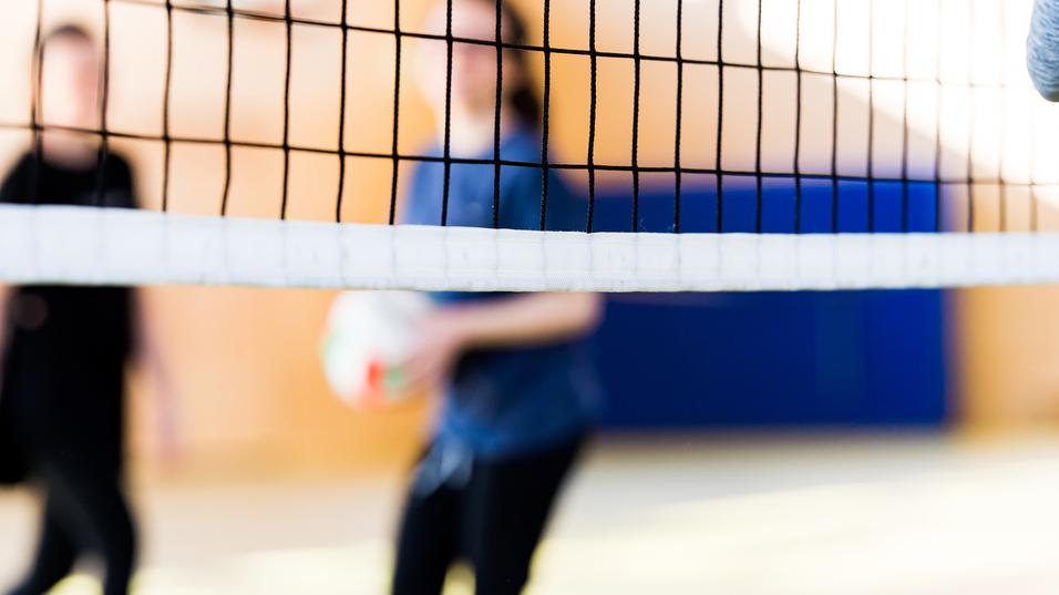 Unscharfe Silhouetten von zwei Personen hinter einem Volleyballnetz in einer Sporthalle. Eine Person hält einen Volleyball.