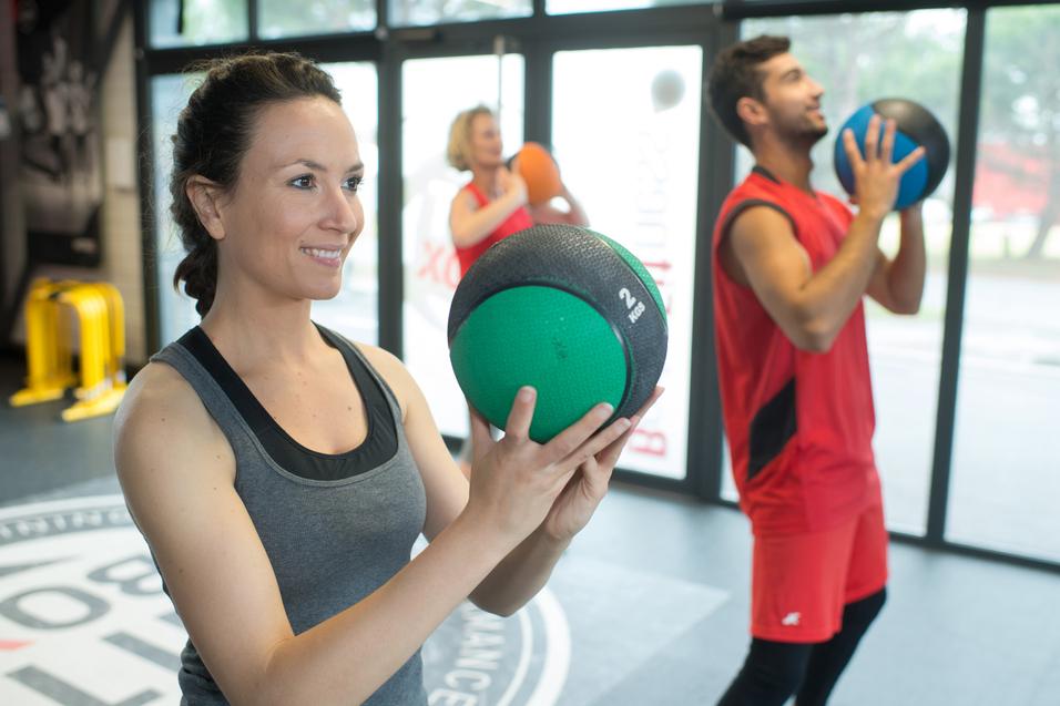 Drei Personen trainieren im Fitnessstudio mit Trainingsbällen; eine Frau hält einen grünen Medizinball.
