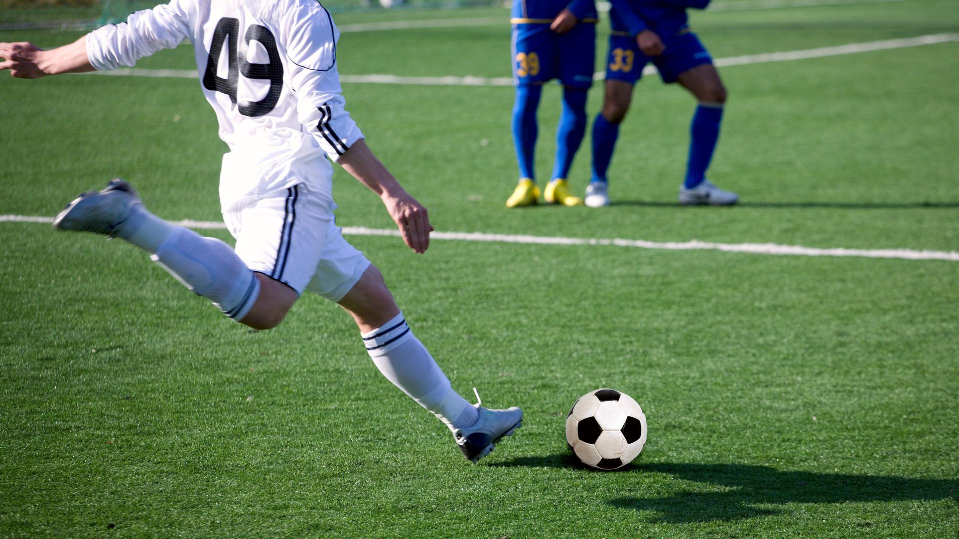 Spieler in weißer Ausrüstung kickt einen Fußball auf einem grünen Kunstrasenplatz, andere Spieler im Hintergrund.