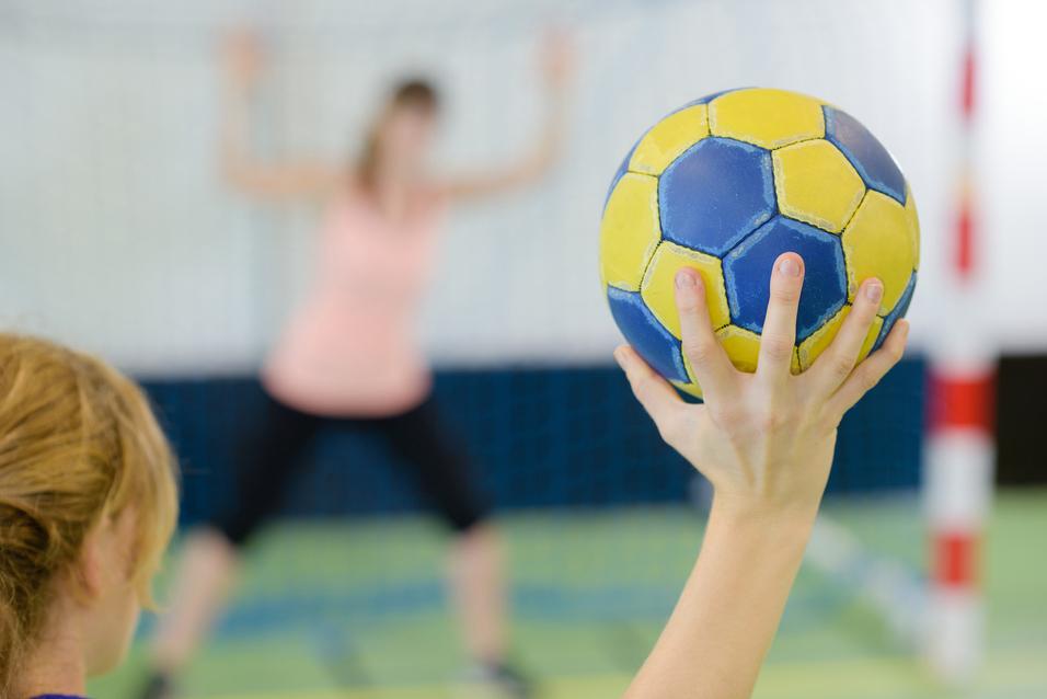 Nahaufnahme einer Hand, die einen gelb-blauen Handball hält, während eine Spielerin im Hintergrund positioniert ist.
