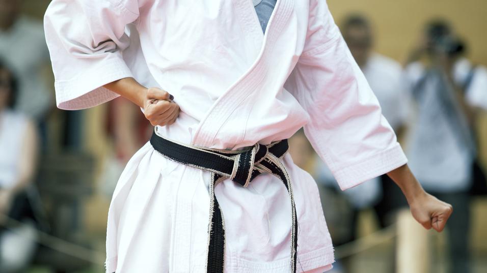 Nahaufnahme eines Karateka in Weiß mit schwarzem Gürtel, der eine Faustballenposition einnimmt.