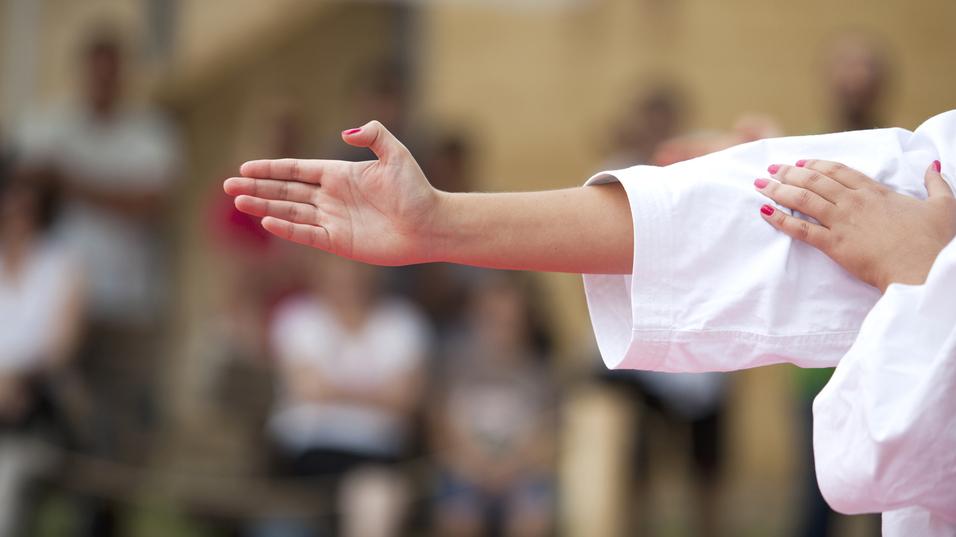 Hand einer Person in Karate-Uniform zeigt eine offene Handfläche, im Hintergrund sind Zuschauer sichtbar.
