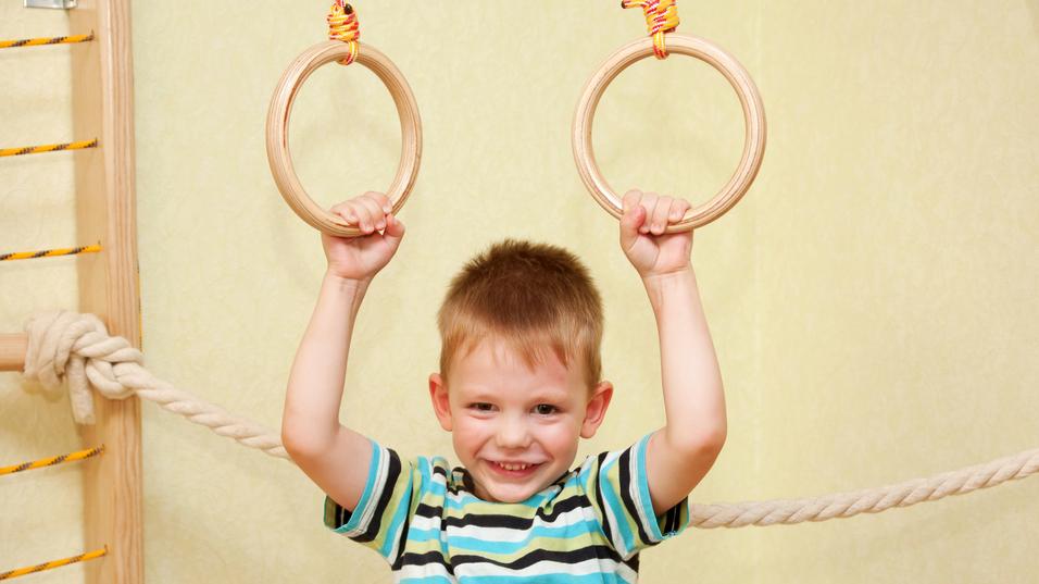 Junge mit einem gestreiften T-Shirt hält sich an zwei Holzringen in einem Spielraum fest und lächelt.
