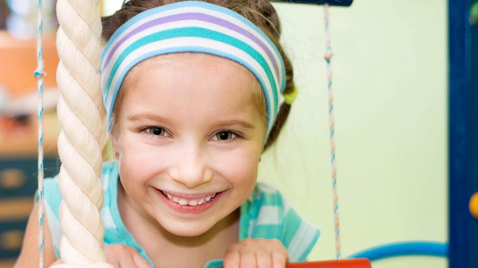 Lächelndes Mädchen mit blauer Kopfband und gestreifter Bluse hält sich an einem Klettergerüst fest.