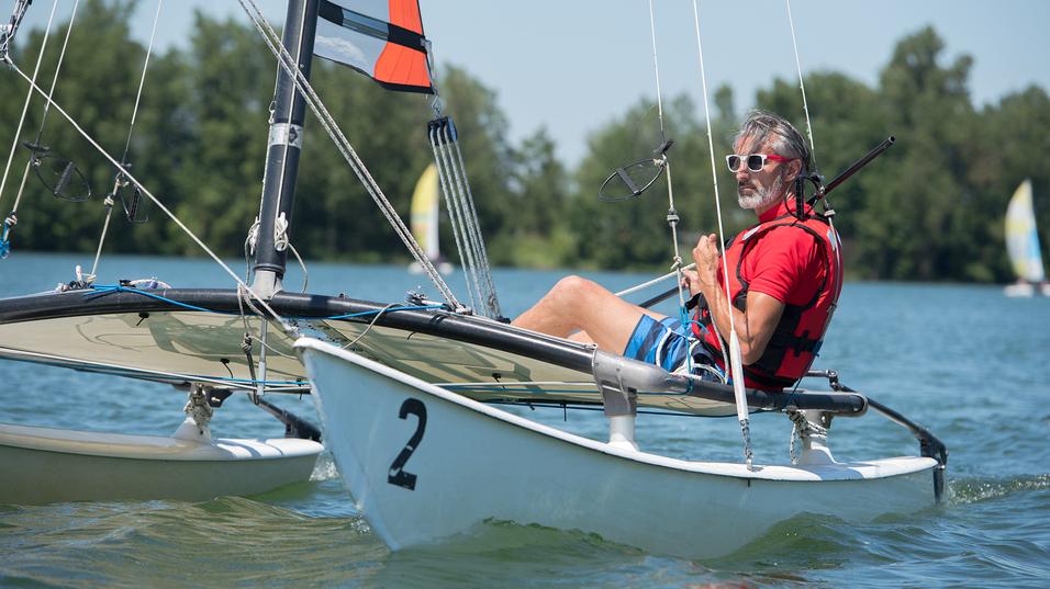 Mann segelt in einem Katamaran auf einem Gewässer, trägt eine Schwimmweste und Sonnenbrille, mit Bäumen im Hintergrund.