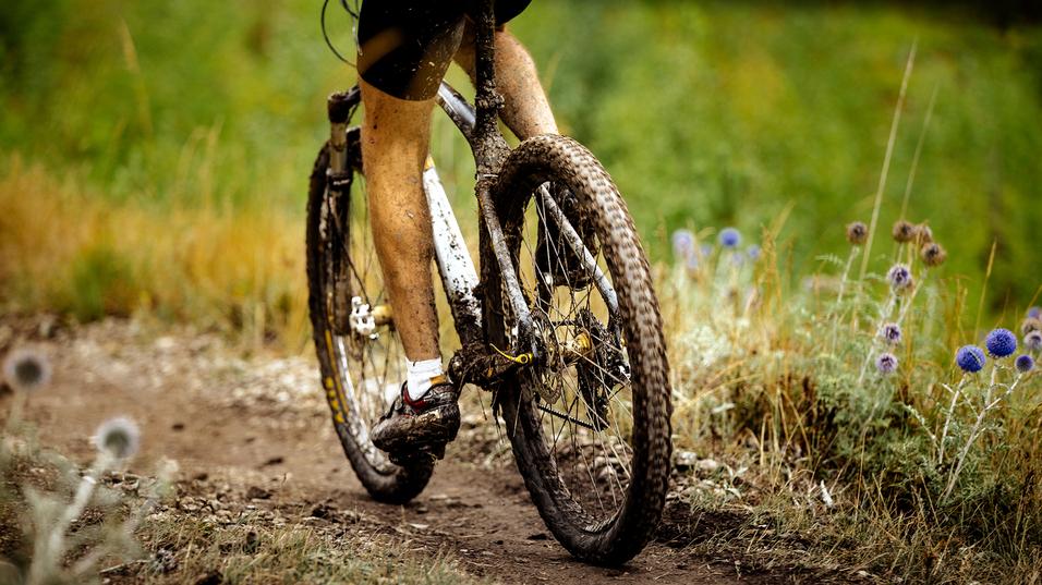 Nahaufnahme eines mountainbikefahrenden Radfahrers mit schmutzigen Beinen auf einem schmalen, unbefestigten Weg.