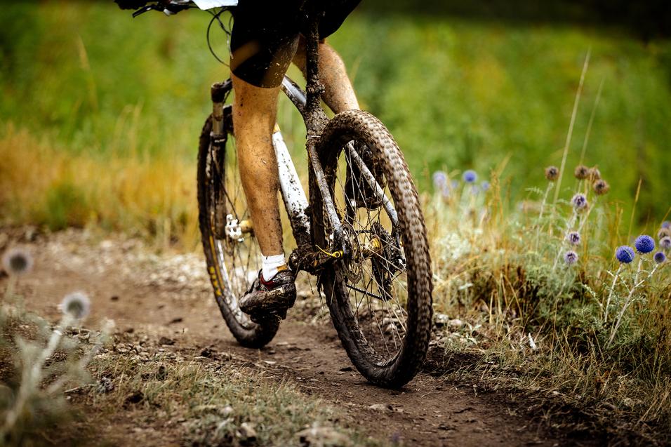 Nahaufnahme eines mountainbikefahrenden Radfahrers mit schmutzigen Beinen auf einem schmalen, unbefestigten Weg.