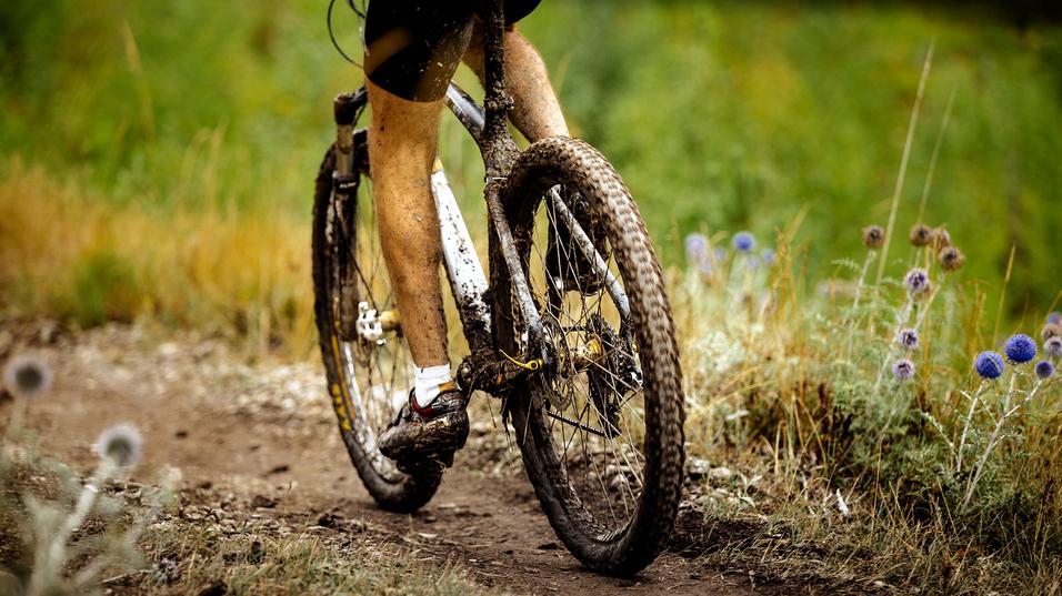 Nahaufnahme eines mountainbikefahrenden Radfahrers mit schmutzigen Beinen auf einem schmalen, unbefestigten Weg.